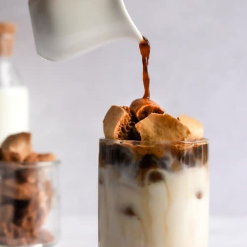 dalgona latte on a table with a hand pouring coffee on top.
