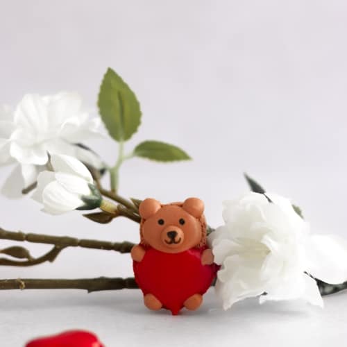 cute macarons in the shape of a bear holding a heart filled with chocolate ganache on a table with a flower branch.