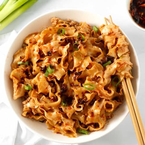 garlic chilli oil noodles in a white bowl with some scallions on the side and a bowl of chilli oil.