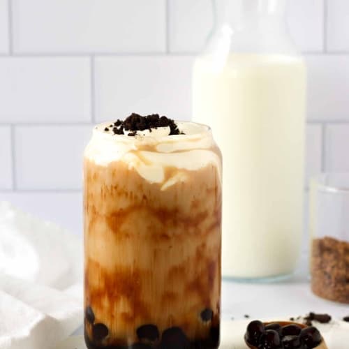 a glass with some milk tea and boba topped with cheese foam and crushed oreo cookies.