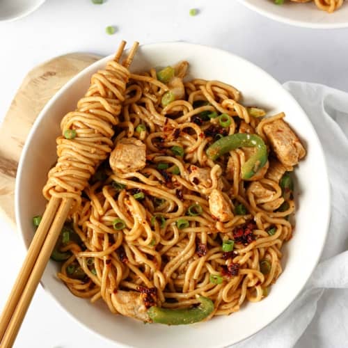 a white bowl with thick wheat noodles coated with a peanut butter chilli oil sauce