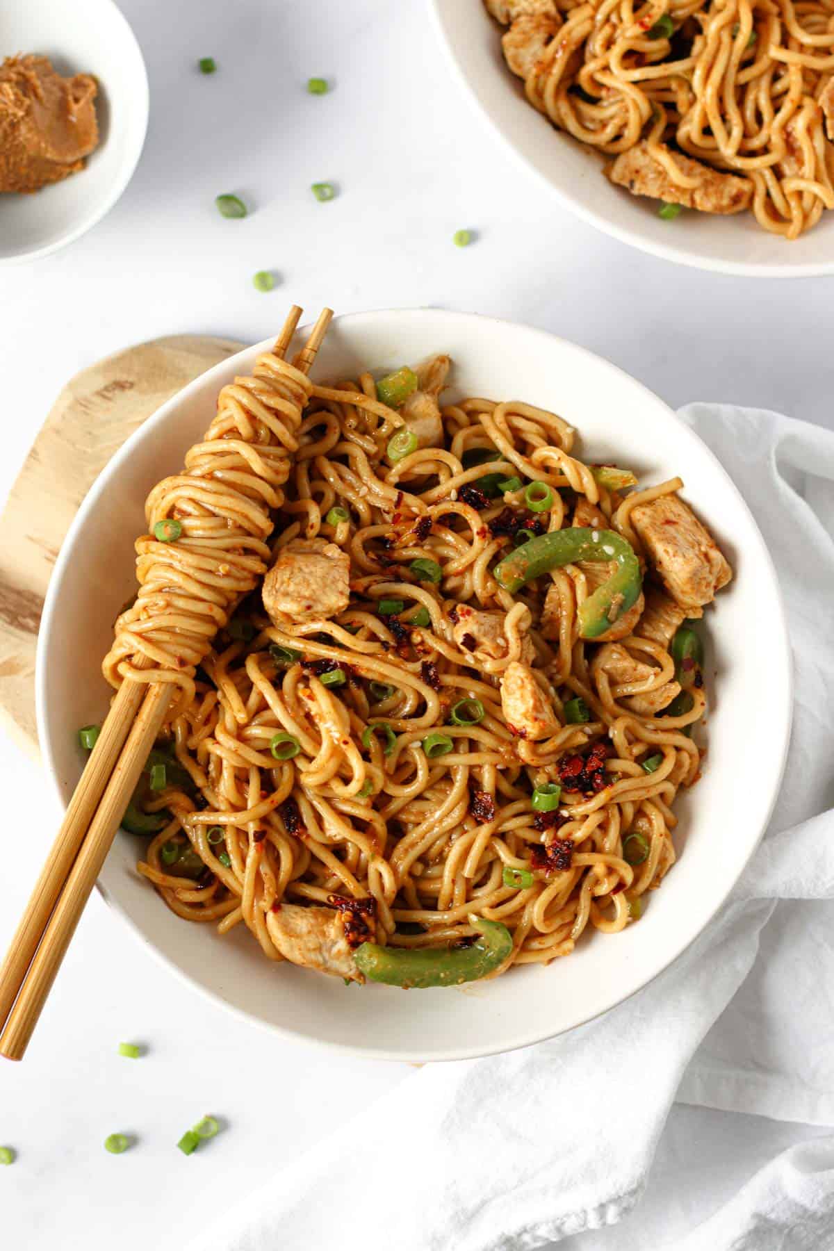 a white bowl with thick wheat noodles coated with a peanut butter chilli oil sauce