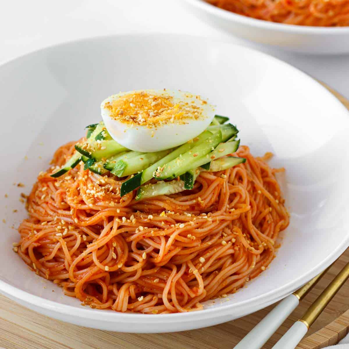 bibim guksu in a bowl featuring somyeon noodles mixed in a gochujang sauce and topped with cucumber, egg and sesame seeds.
